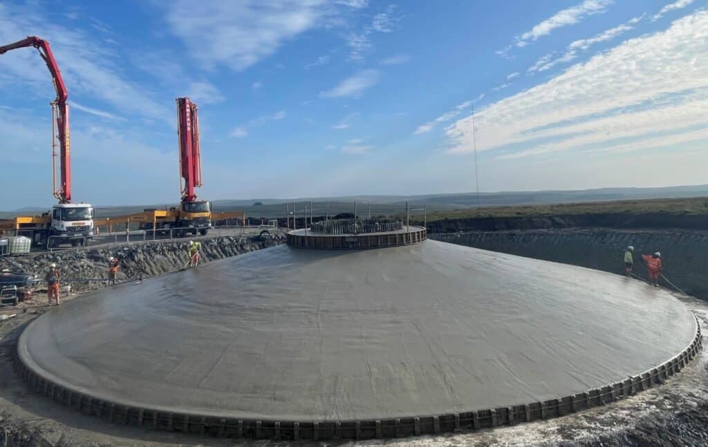 A circular reinforced concrete foundation under construction at dusk, surrounded by rocky ground and equipment, with hills and low-lying mist in the background beneath a darkening sky.